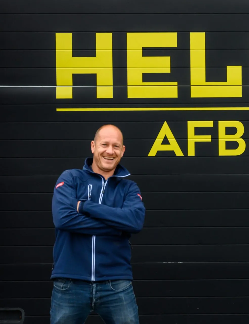 Ruud van den Akker, eigenaar van Helmex Afbouw, met de armen over elkaar voor de zwarte gevel met het gele Helmex-logo